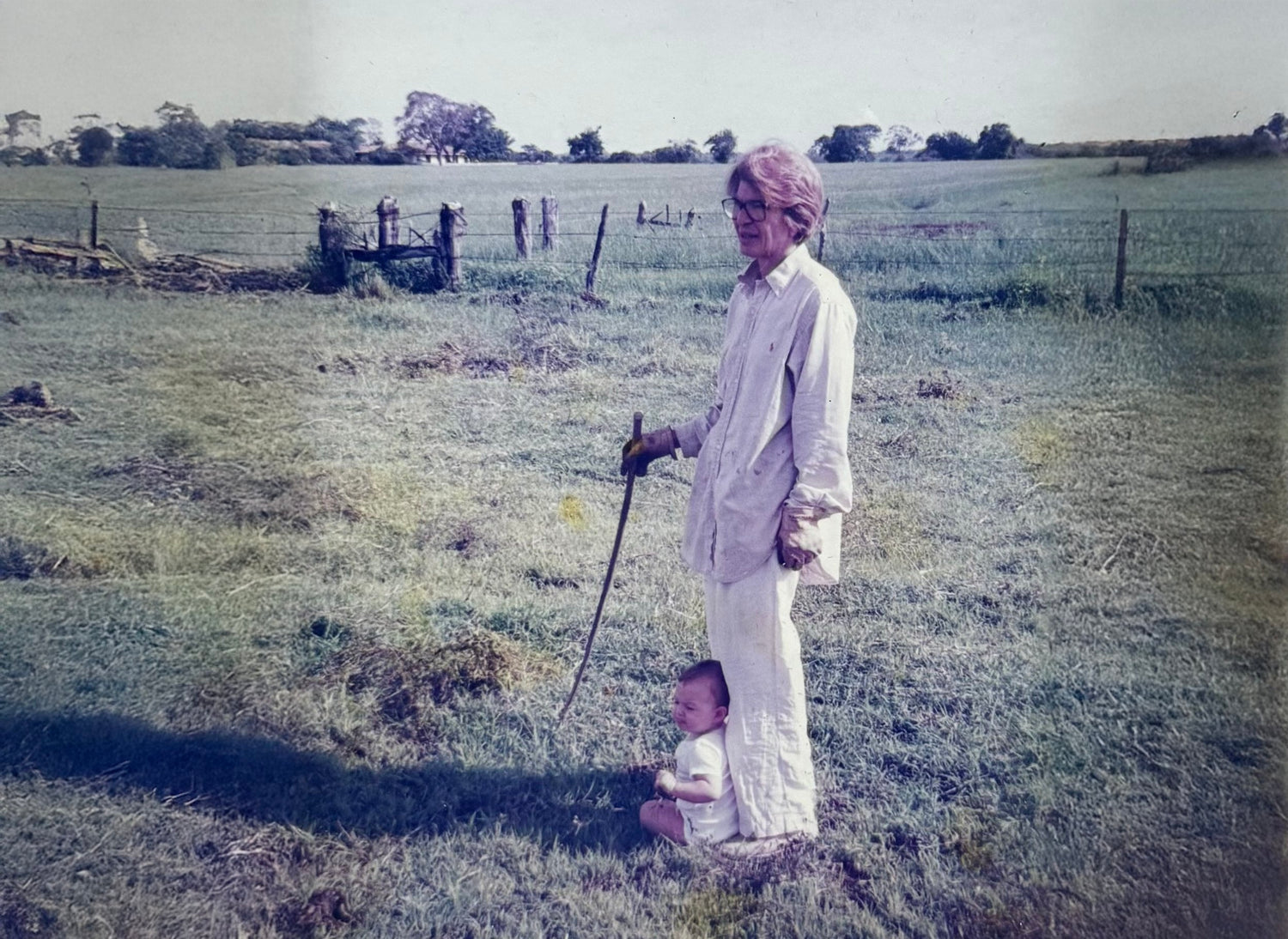 An elderly woman with glasses, dressed in light-colored clothing, standing outdoors in a grassy field, holding a rake, with a small baby sitting at her feet.