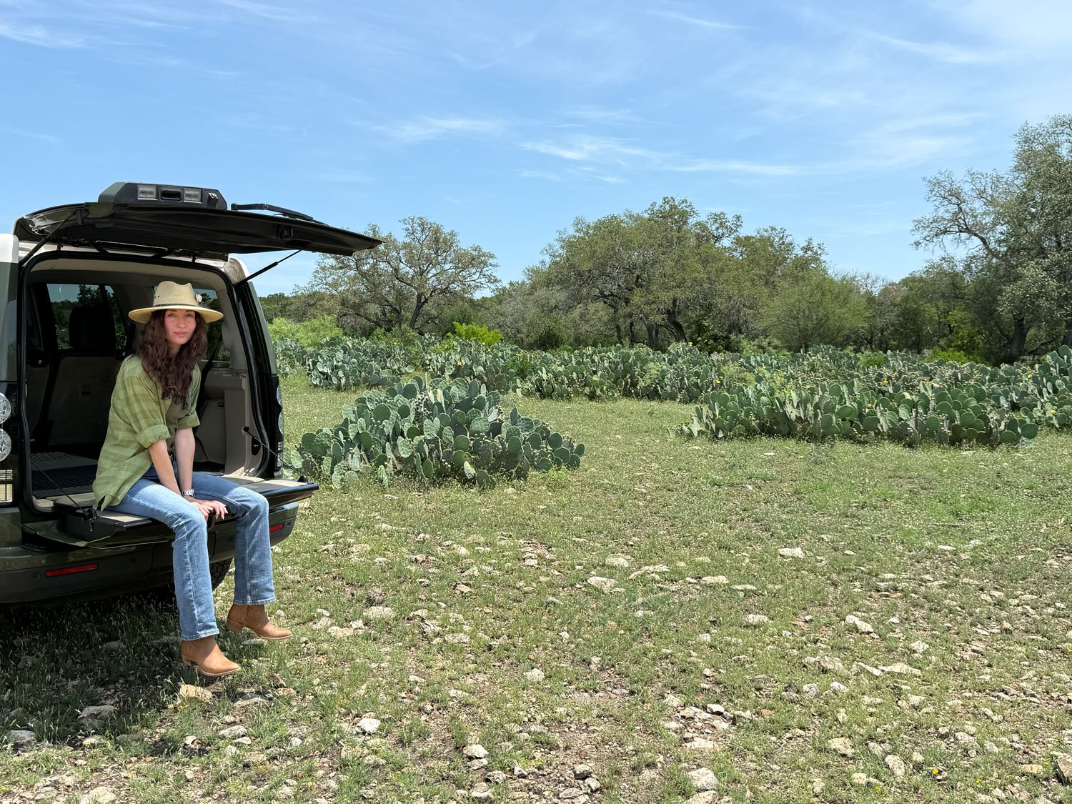 A woman in a green plaid shirt and jeans sitting on the open trunk of a black SUV in a desert landscape with cactus plants and trees under a partly cloudy sky.